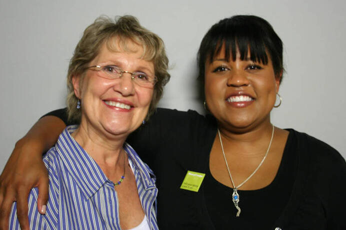 Older white woman posing with younger Black woman, both smiling, against light backdrop