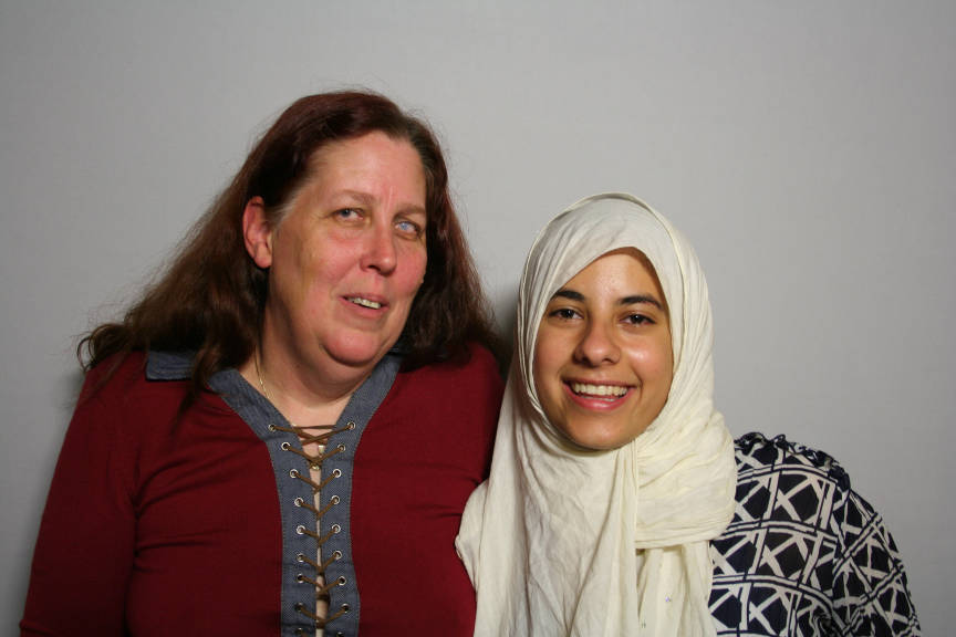 Two women smile for the camera, one with a red lace-up shirt and long brown hair, and the other in a black and white shirt and wearing a headscarf